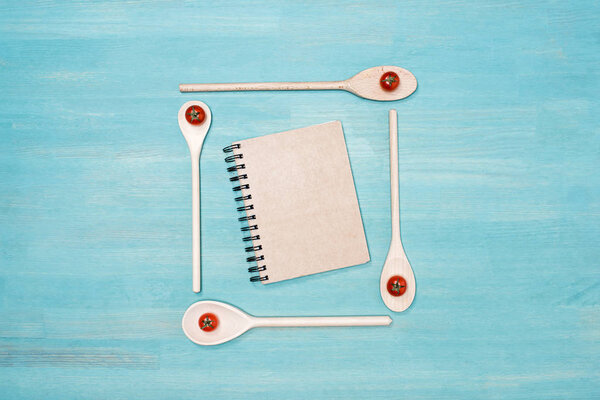 Top view of wooden spoons with cherry tomatoes and closed cookbook with blank cover on wooden table