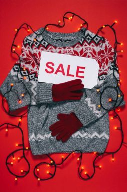 top view of winter sweater, mittens, price tag with sale lettering and christmas lights isolated on red clipart