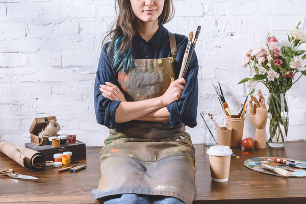 cropped image of female artist sitting on table with crossed arms and holding brushes 