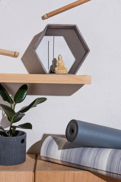 small statue of buddha in frame with incense stick on wooden shelf, yoga mats near plant