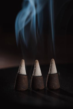 three burning incense sticks with smoke on dark surface clipart