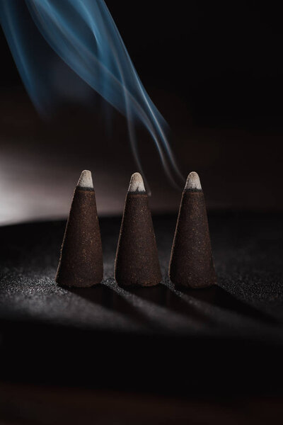 three burning incense sticks with blue smoke on black