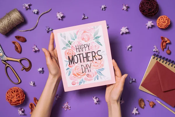 部分视图的妇女举行快乐的母亲节明信片在手上的装饰品周围的紫色隔离 — 图库照片