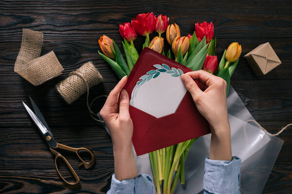 cropped shot of woman with postcard in hands and tulips in wrapping paper on wooden tabletop