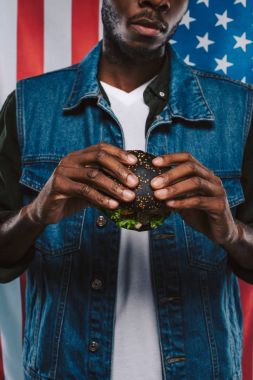 cropped shot of african american man holding black burger against usa flag clipart