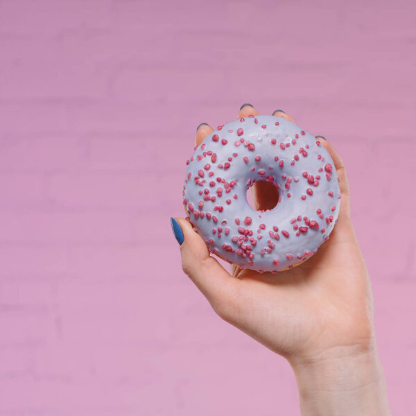 обрезанный снимок женщины, держащей сладкий глазурованный пончик в руке перед розовой кирпичной стеной
