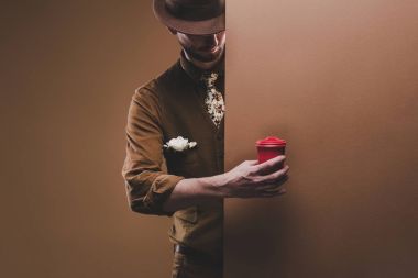 Stylish man in hat holding coffee in red paper cup isolated on brown clipart