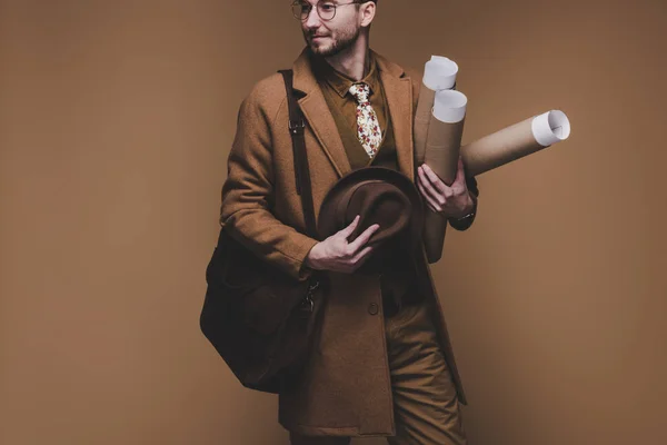 Young man in vintage styled clothes holding rolled papers isolated on brown
