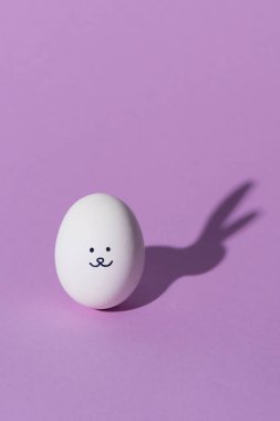 one chicken egg with smiley and rabbit shadow on purple surface clipart