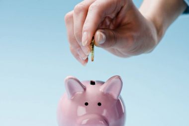 partial view of woman putting coin into pink piggy bank isolated on blue clipart