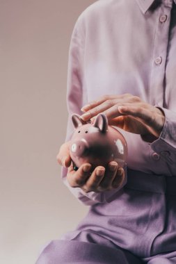 partial view of woman holding pink piggy bank in hands isolated on lilac clipart