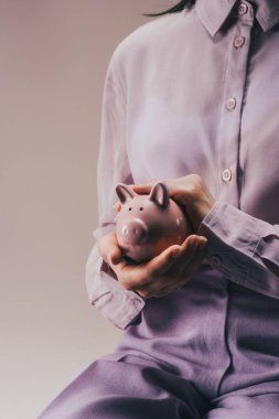 partial view of woman holding pink piggy bank in hands isolated on lilac clipart