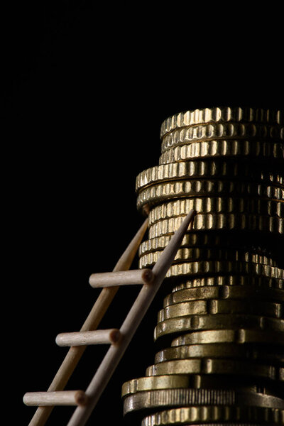 close up view of pile of coins with little wooden ladder isolated on black