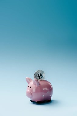 Close up view of pink piggy bank and silver bitcoin on blue backdrop clipart