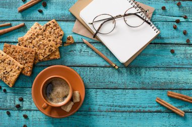 flat lay with cup of coffee, cookies, eyeglasses, empty notebook, roasted coffee beans and cinnamon sticks around on blue wooden tabletop clipart