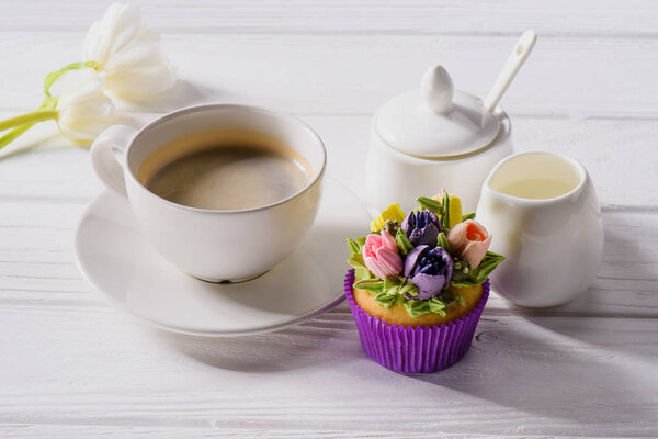 close up view of tulips, cup of coffee, sweet muffin and jag of cream on white wooden tabletop