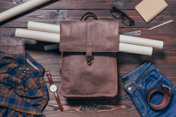 flat lay with male shirt, jeans, watch and blueprints in backpack arranged on wooden tabletop