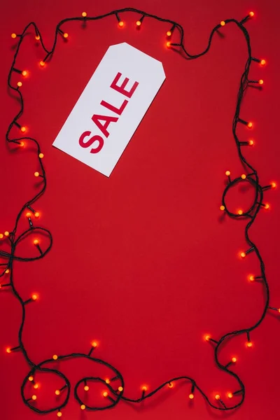 Flat lay with price tag with sale lettering and christmas lights isolated on red — Stock Photo