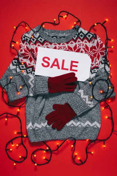 Top view of winter sweater, mittens, price tag with sale lettering and christmas lights isolated on red — Stock Photo