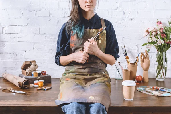 Cropped image of female artist sitting on table and holding brushes in workshop — Stock Photo