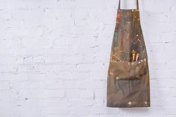 Brown apron with paint brushes hanging on white brick wall — Stock Photo