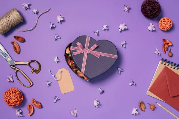 Plano con caja de regalo en forma de corazón, flores, tijeras, cuerda y sobre aislado en púrpura - foto de stock