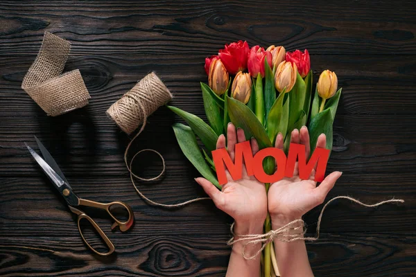 Vue partielle de la femme avec lettrage maman dans les mains et bouquet de tulipes sur table en bois, concept de fête des mères — Photo de stock