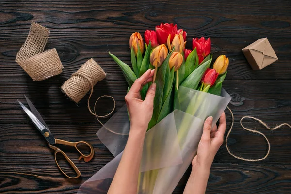 Vista parziale di donna che avvolge mazzo di tulipani in carta da imballaggio su tavolo in legno — Foto stock