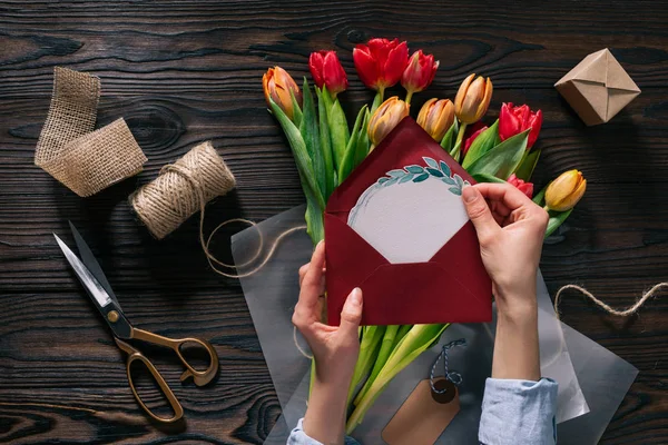 Cropped shot of woman with postcard in hands and tulips in wrapping paper on wooden tabletop — Stock Photo