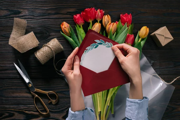 Plan recadré de femme avec carte postale dans les mains et tulipes en papier d'emballage sur table en bois — Photo de stock