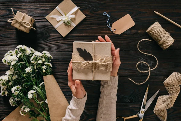 Vista parcial de la mujer sosteniendo regalo con flores y suministros de decoración en la mesa de madera - foto de stock