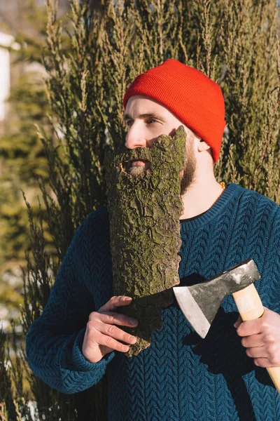Hombre con barba hecha de corteza de madera y hacha en la mano mirando al aire libre - foto de stock