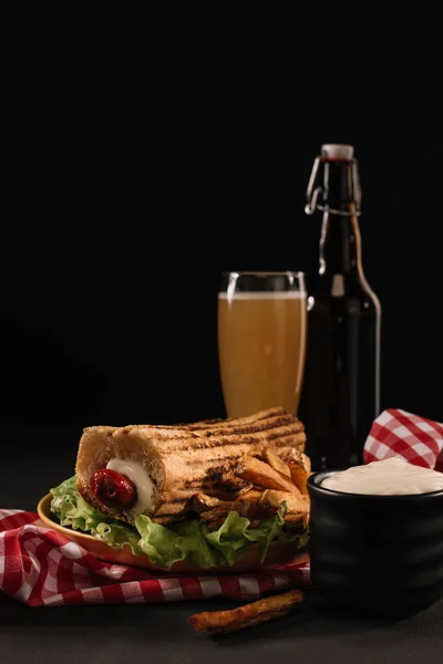 Délicieux hot dog français sur assiette avec bouteille et verre de bière isolé sur noir — Photo de stock