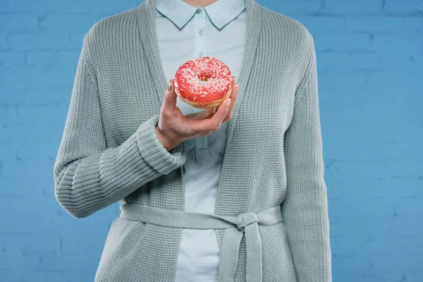 Cropped shot of woman in sweater and shirt holding delicious glazed doughnut in front of blue brick wall — Stock Photo