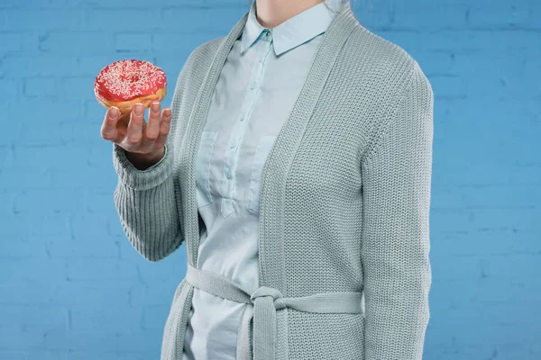 Обрезанный снимок женщины в свитере и рубашке с вкусным глазурованным пончиком перед синей кирпичной стеной — стоковое фото