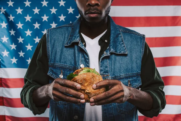 Recortado disparo de hombre afroamericano sosteniendo hamburguesa contra bandera de EE.UU. - foto de stock