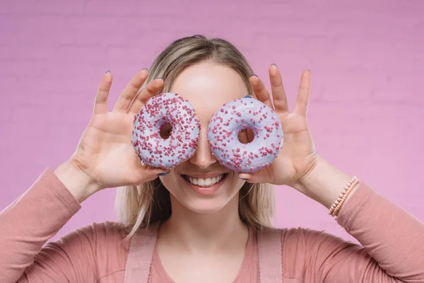 Mujer joven sonriente cubriendo los ojos con donuts frente a la pared de ladrillo rosa - foto de stock