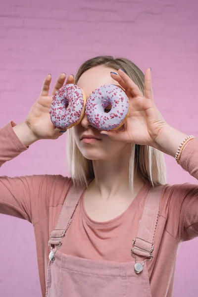 Giovane donna che copre gli occhi con ciambelle davanti al muro di mattoni rosa — Foto stock