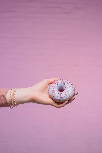 Tiro cortado de mulher segurando donut envidraçado na mão na frente da parede de tijolo rosa — Fotografia de Stock
