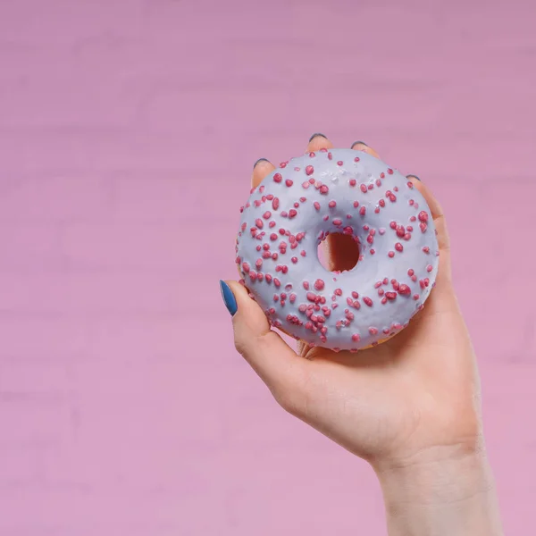 Tiro recortado de la mujer sosteniendo rosquilla acristalada dulce en la mano delante de la pared de ladrillo rosa - foto de stock