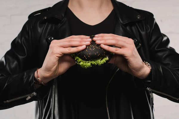 Ritagliato colpo di uomo in giacca di pelle in possesso di hamburger nero — Foto stock