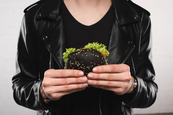 Recortado tiro de hombre en elegante chaqueta de cuero sosteniendo hamburguesa negro - foto de stock