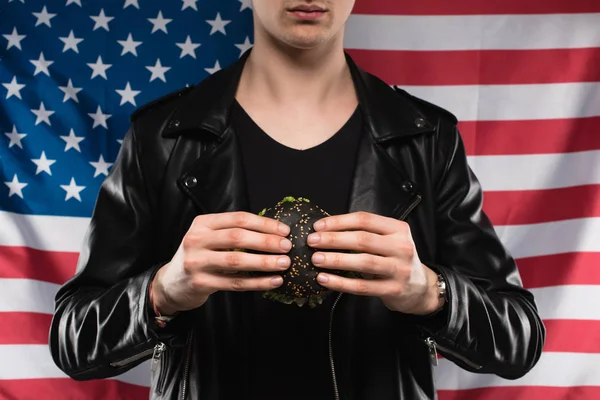 Recortado disparo de hombre joven en chaqueta de cuero sosteniendo hamburguesa negra contra bandera de EE.UU. - foto de stock