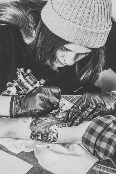 Foto en blanco y negro de la mujer artista del tatuaje en guantes trabajando en la pieza del brazo - foto de stock