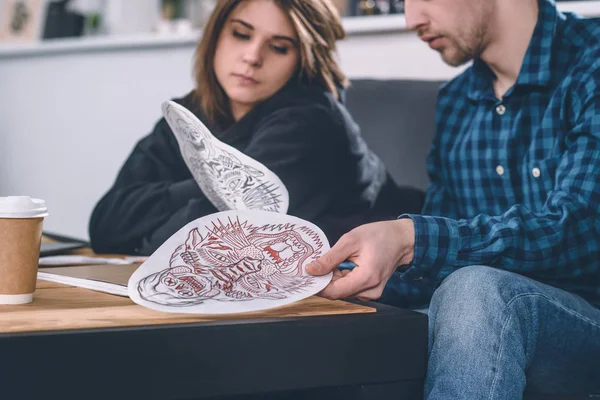 Different tattoo designs in hands of young couple in tattoo salon — Stock Photo