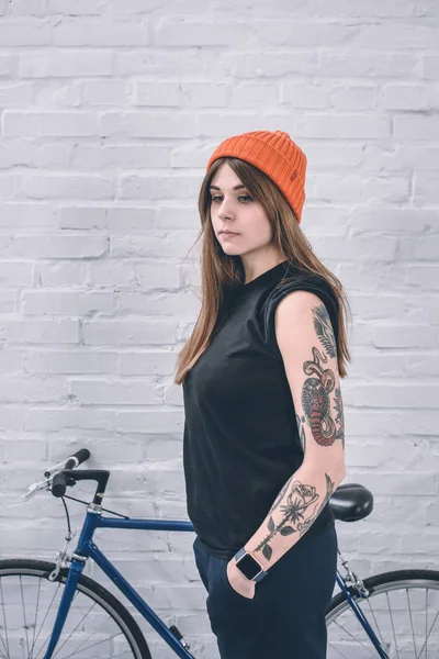Chica con estilo con el brazo tatuado de pie junto a la bicicleta por la pared - foto de stock