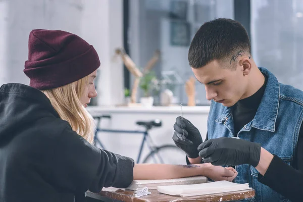 Artista del tatuaje en guantes trabajando en pieza de brazo femenino en estudio - foto de stock