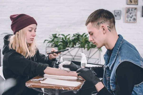 Young woman taking photo of tattooing process in studio — Stock Photo