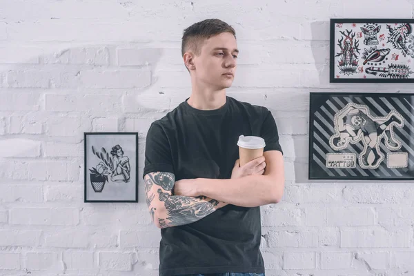 Hombre tatuado con taza de café junto a la pared en el estudio de tatuajes - foto de stock