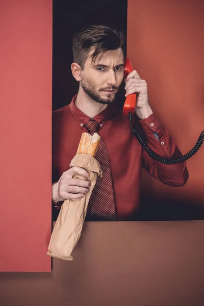 Hombre reflexivo sosteniendo baguette y auriculares del teléfono por paredes de papel rojo aisladas en negro - foto de stock